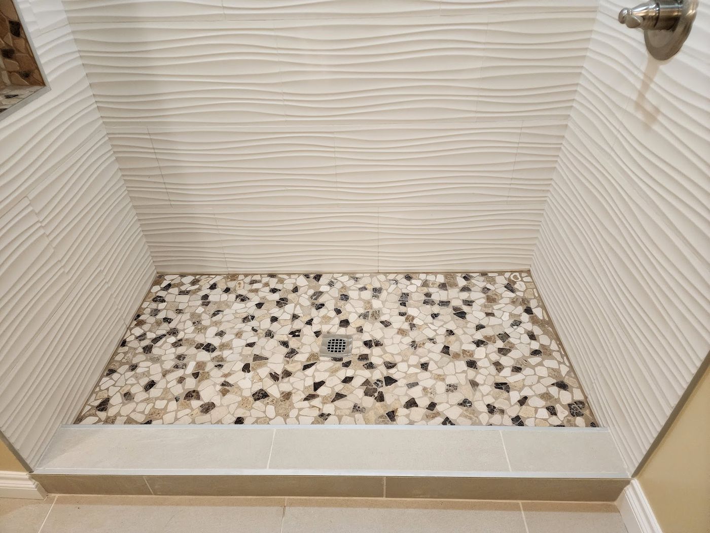 A walk-in shower with rippled white wall tile and a pebble mosaic floor in cream, tan and black, with a stone threshold.